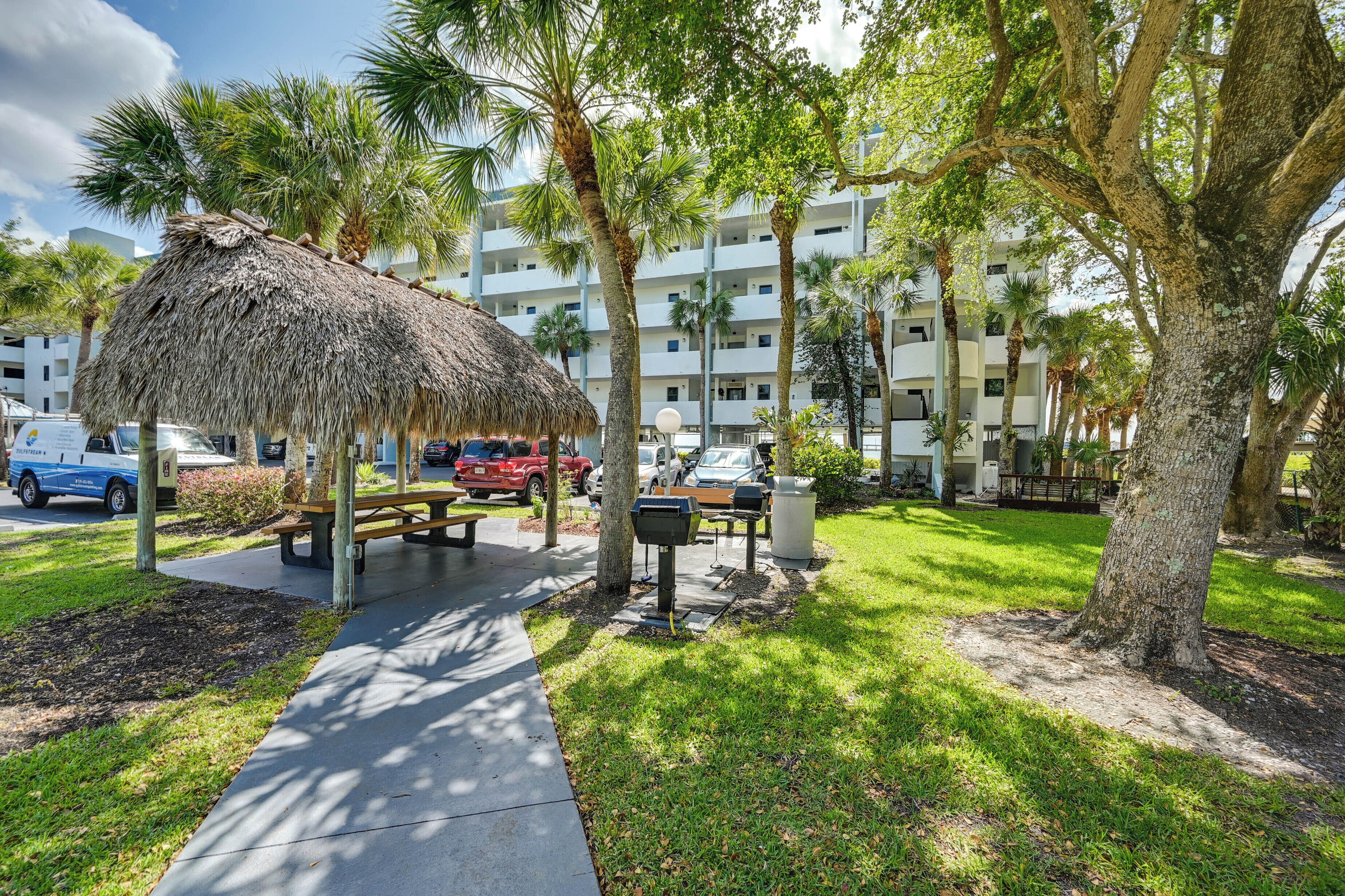 A thatched-roof pavilion with a picnic table, surrounded by palm trees and a multi-story building in the background.