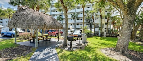 A thatched-roof pavilion with a picnic table, surrounded by palm trees and a multi-story building in the background.