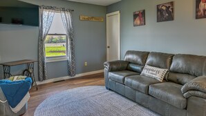A living room with a leather sofa, a blue chair, a grey rug, a window with curtains, and wall art. - Upstate New York Vacation Rental Near Cooperstown! (Jordanville)