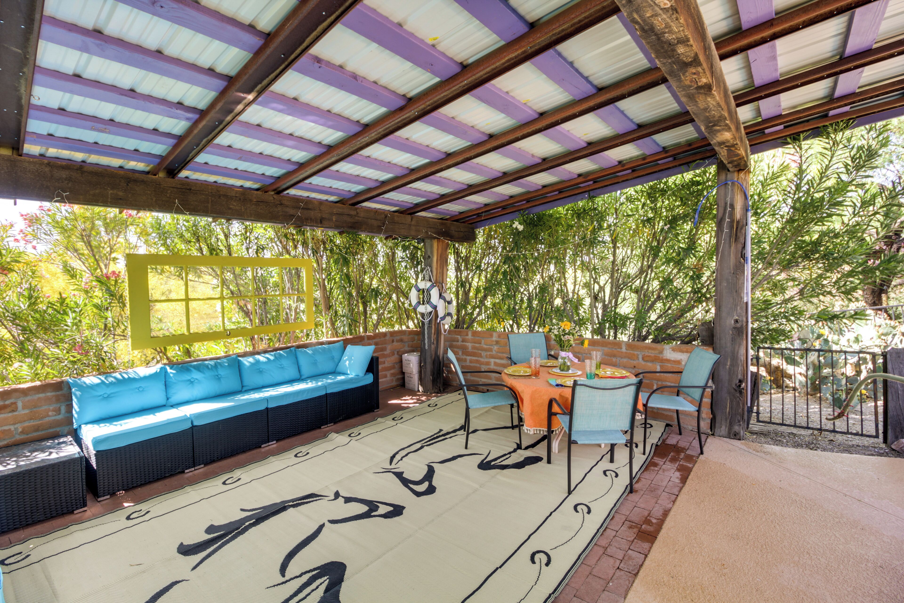 A covered patio with a blue sofa, a dining table with chairs, and a view of greenery.