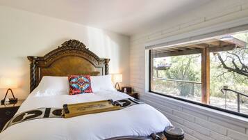 A neatly made bed with a wooden headboard, white linens, and a colorful pillow. There is a wooden tray on the bed, and a window with a view of trees and a patio outside.