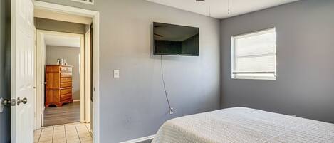 Een slaapkamer met een bed, een plafondventilator, een aan de muur bevestigde televisie en een raam met blinds.