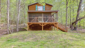 A wooden cabin with a balcony and a staircase leading up to it, surrounded by trees.