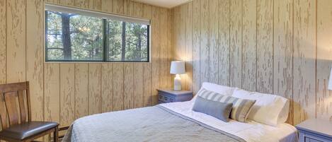 A bedroom with a large bed, a chair, a nightstand with a lamp, and a window with a view of trees.