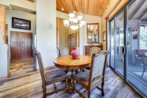 A dining area with a wooden table and chairs, a chandelier, and a large window with a view. - Luxe Resort Cabin w/ Deck: Hike, Swim, Relax! (Show Low)