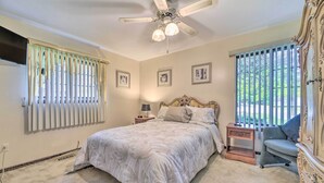 Ein Schlafzimmer mit Bett, Nachttisch, Sessel, Deckenventilator und einem Fenster mit Jalousien.