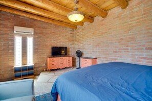 Een slaapkamer met een bakstenen muur, houten balken in het plafond, een bed, een dressoir, een televisie en een ventilator.