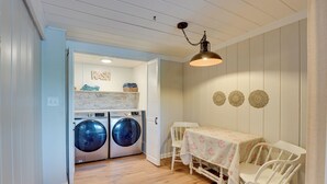 A laundry room with two front-loading washing machines, a table with a floral tablecloth, and a white chair. - Big Pine Island Lake Cottage w/ Boat Dock & Kayaks (Belding)