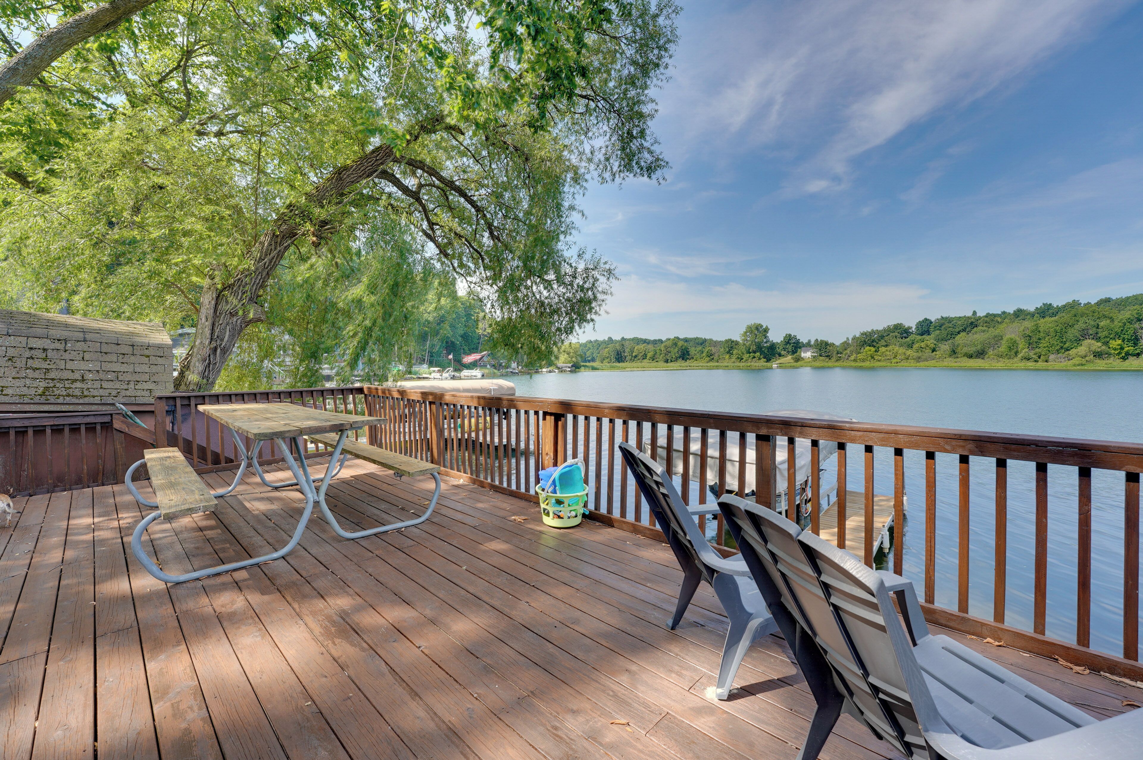 A wooden deck with picnic tables and chairs overlooking a calm lake, surrounded by trees.