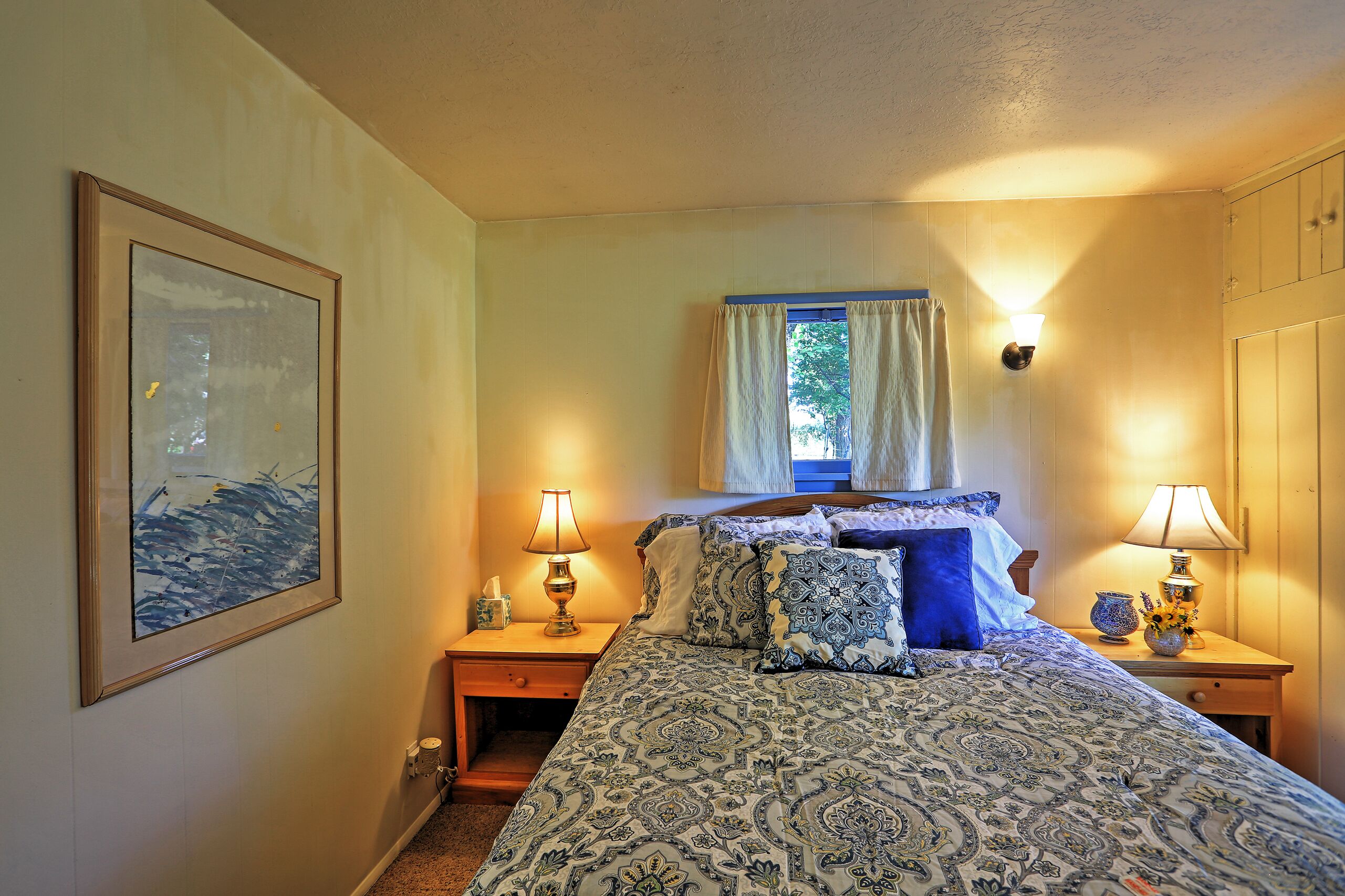 A bedroom with a bed, two bedside tables, a nightstand, a framed picture, a window with curtains, and a wall-mounted lamp.