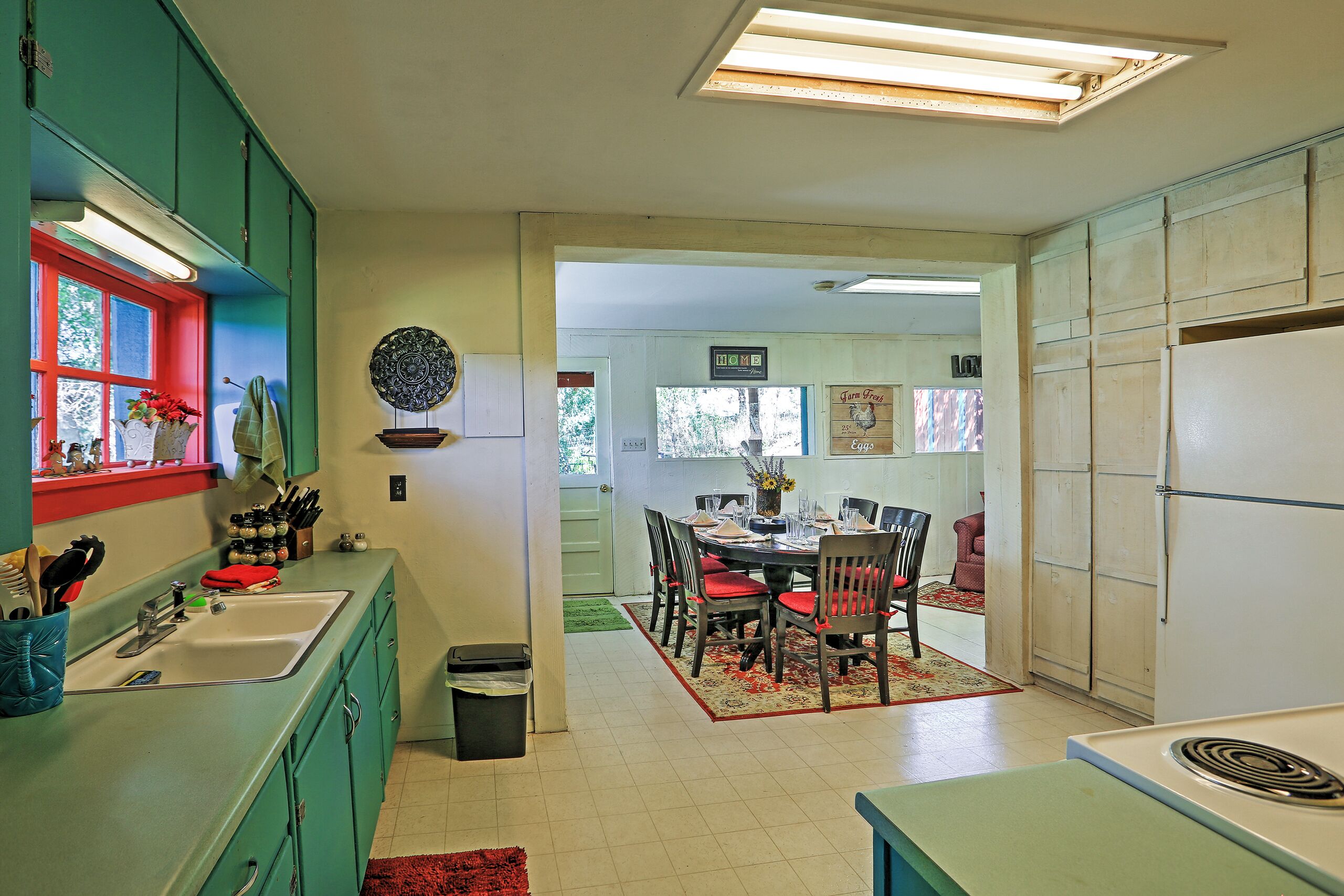 Una cocina con gabinetes verde mar, un ventanal rojo y una zona de comedor con una mesa y sillas.