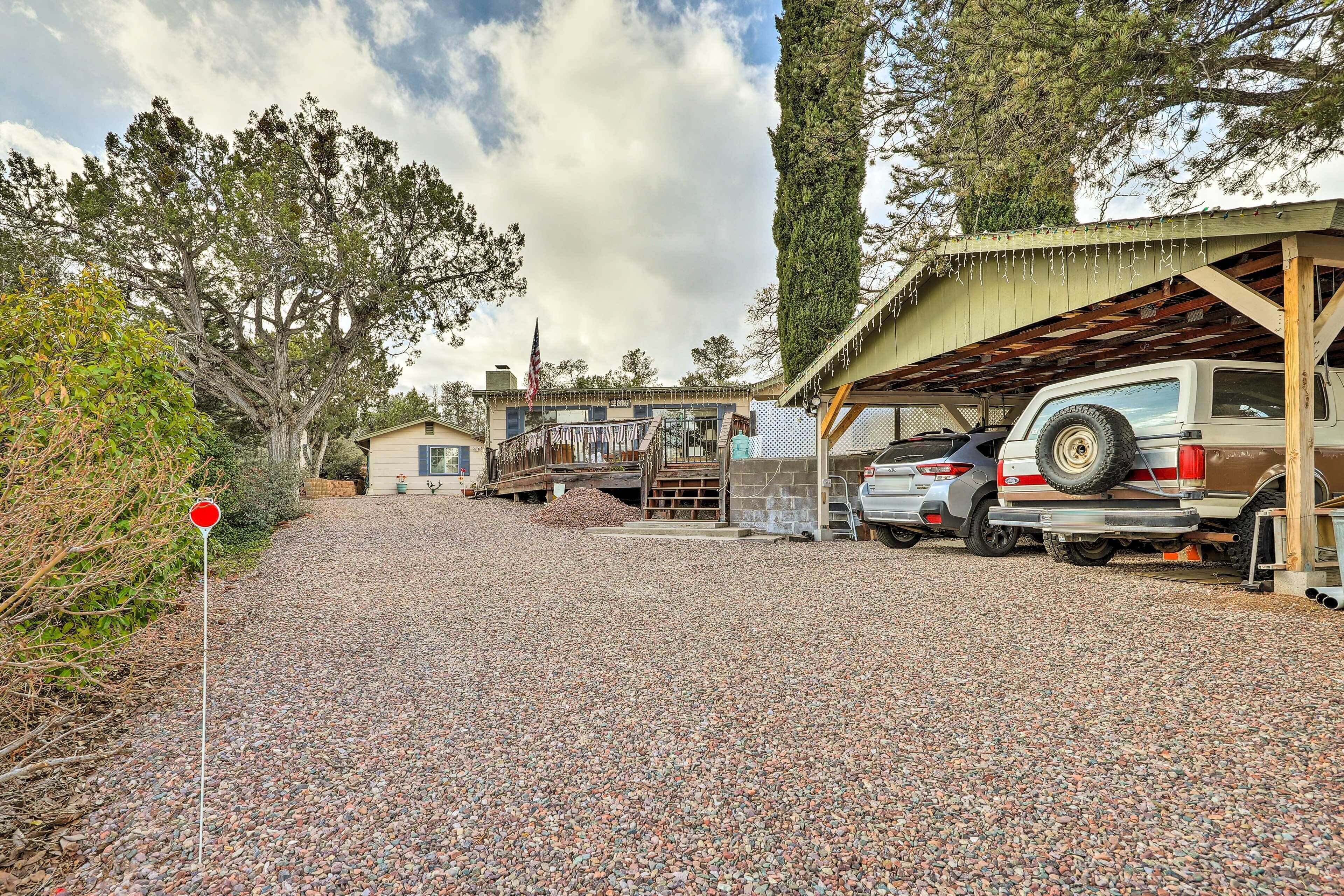 Une allée de gravier mène à un espace de stationnement couvert où se trouve un véhicule, ainsi qu'à un bâtiment doté d'une terrasse en bois.