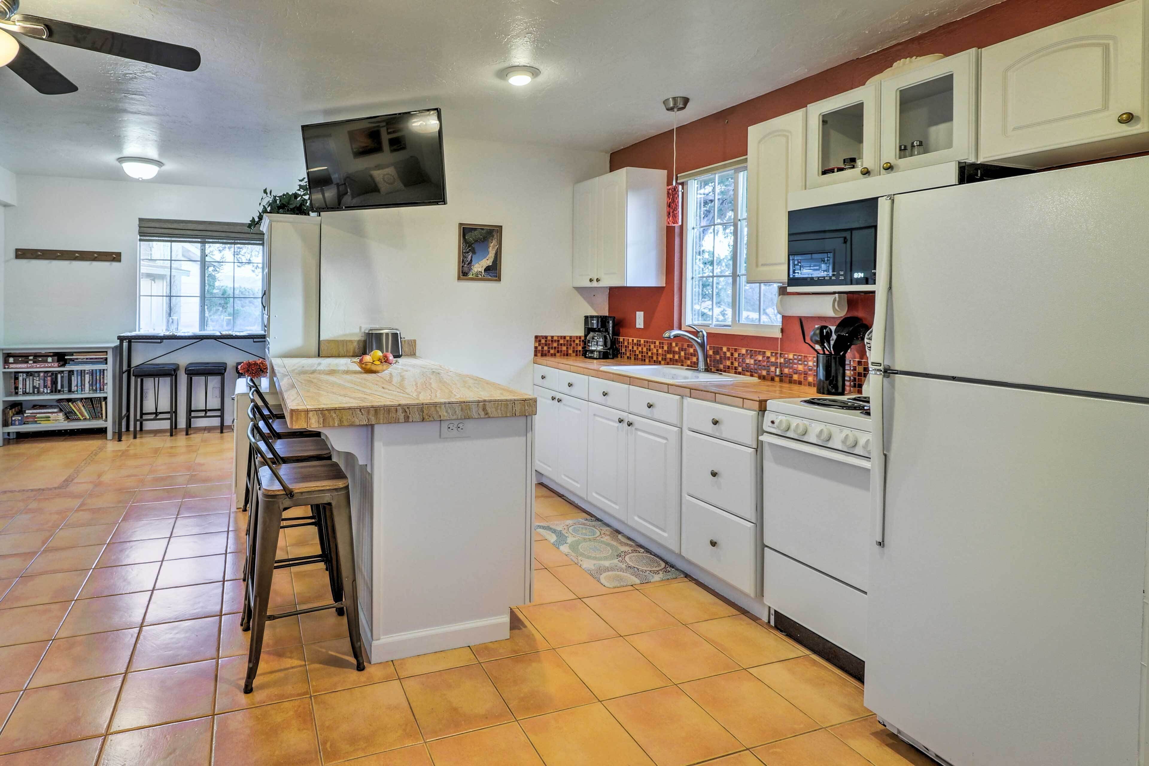 A kitchen with white cabinets, a stove, and a refrigerator. There is a window with a view of trees, and a ceiling fan.