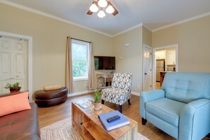 A living room with a brown leather sofa, a blue armchair, a wooden coffee table with a remote and a book, a patterned armchair, a television, and a kitchen visible through an open door. - Sweet Southern Pines Abode w/ Yard & Covered Porch (Southern Pines)