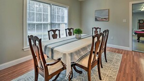 A dining room with a table set for six, surrounded by wooden chairs, a potted plant, and a painting on the wall. - Stokesdale Vacation Rental w/ Game Room! (Stokesdale)
