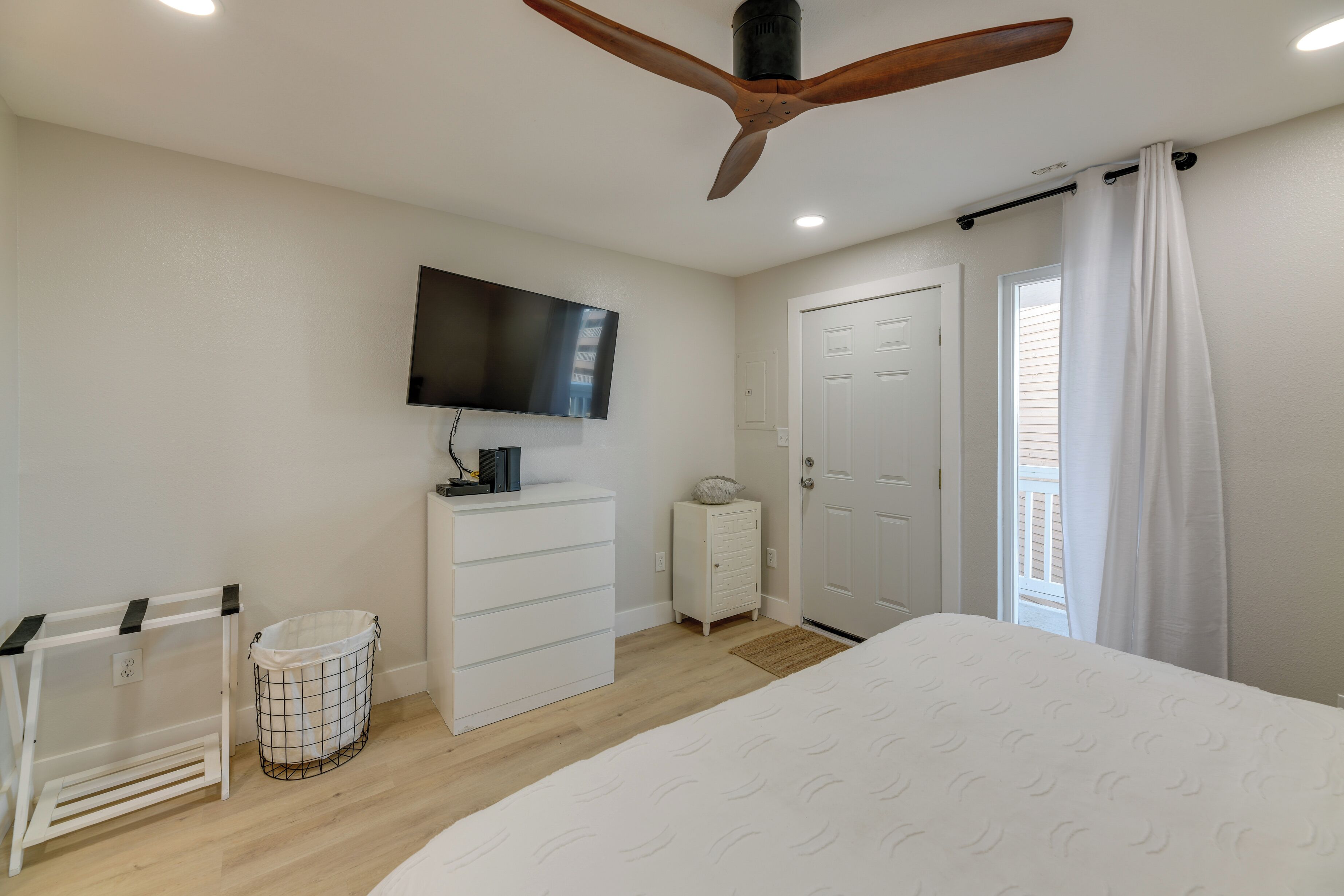 A bedroom with a bed, a ceiling fan, a TV mounted on the wall, a white dresser, a small trash bin, and a door with a curtain.