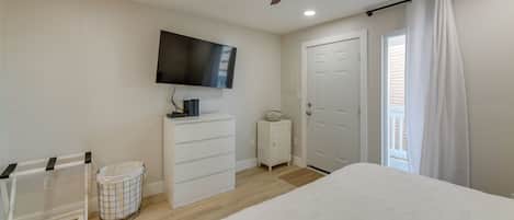 A bedroom with a bed, a ceiling fan, a TV mounted on the wall, a white dresser, a small trash bin, and a door with a curtain.