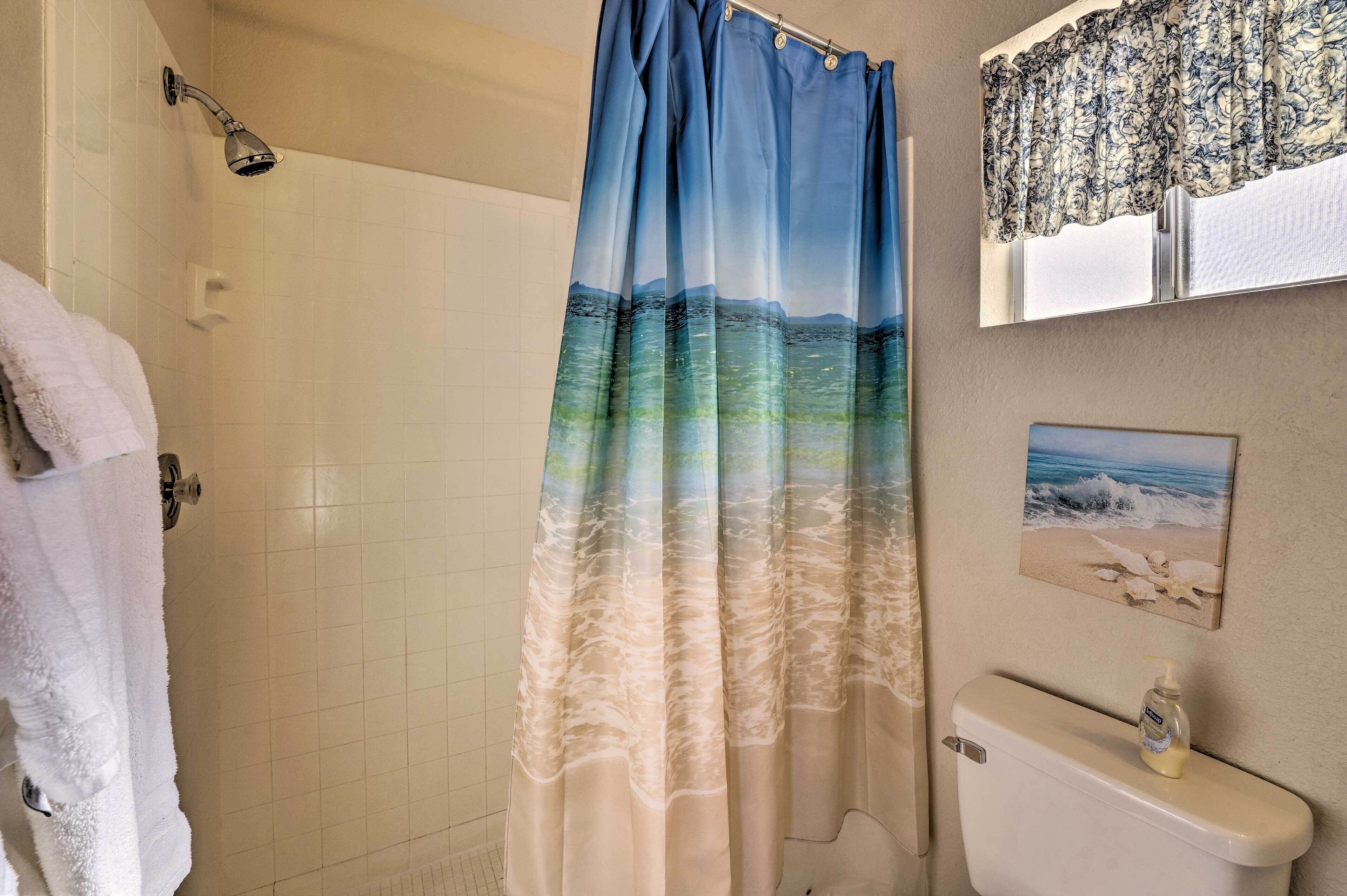 A bathroom with a shower curtain depicting a beach scene, a toilet, a hand soap dispenser, and a framed picture on the wall.