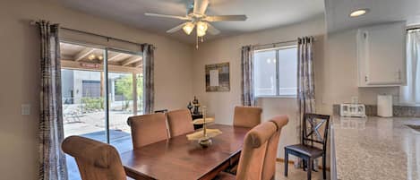 Un coin repas avec une table en bois et six chaises, un ventilateur de plafond et une vue sur l’extérieur, accessible par des portes vitrées.