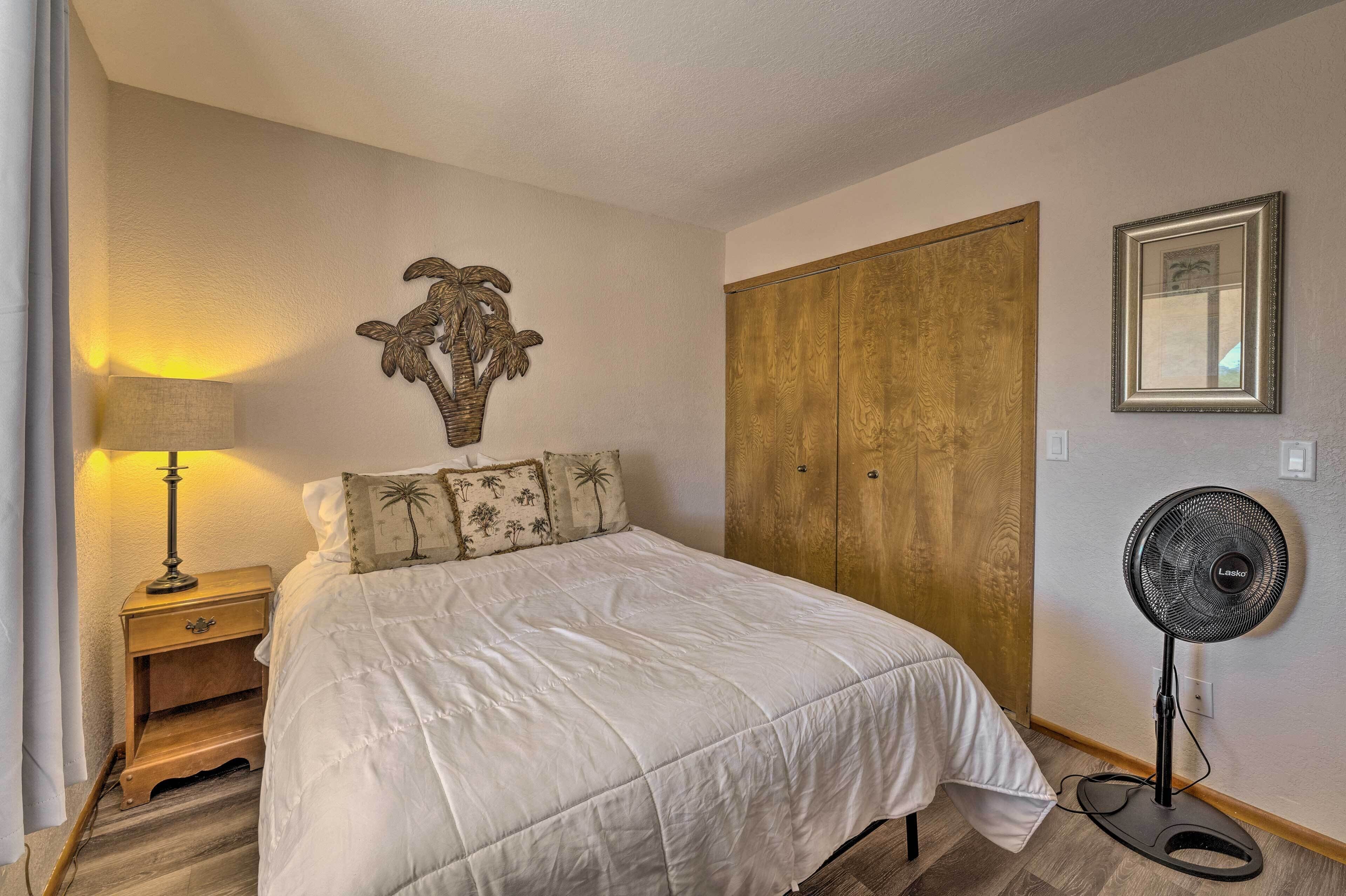 Un dormitorio con cama, mesita de noche, lámpara, decoración en la pared, armario y ventilador de pie.