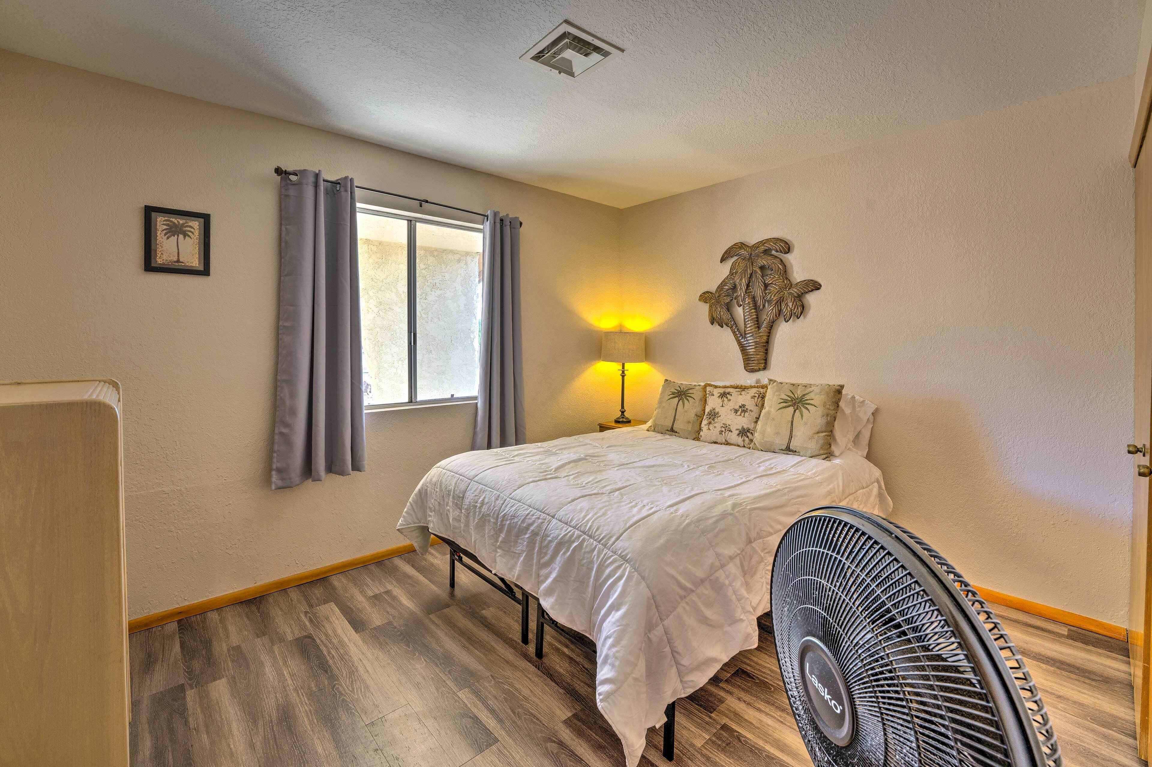 A bedroom with a bed, a standing fan, a window with curtains, a wall-mounted lamp, and a small framed picture.