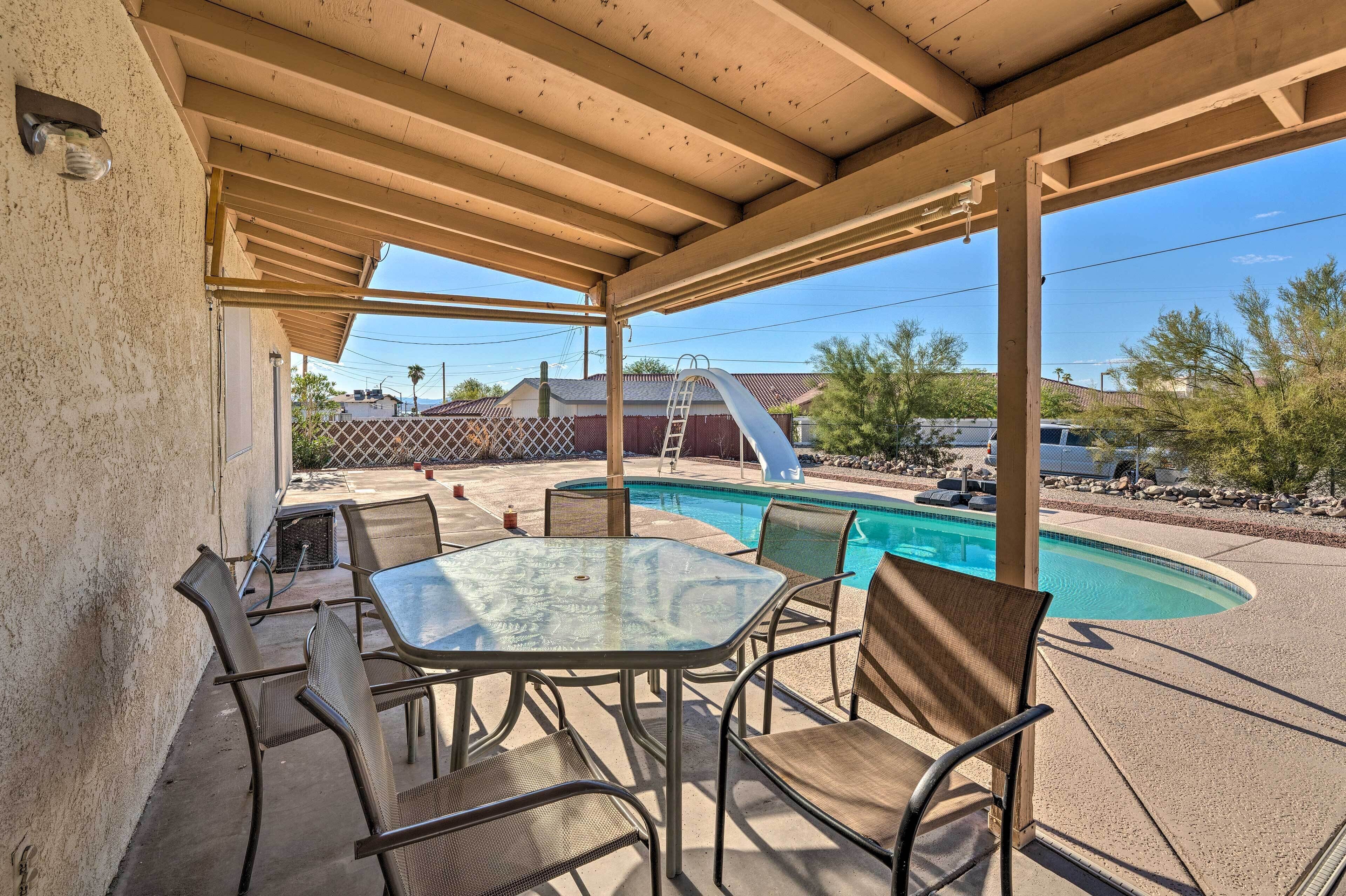Un patio al aire libre con una mesa y sillas de vidrio, una piscina y un área techada con techo de madera.