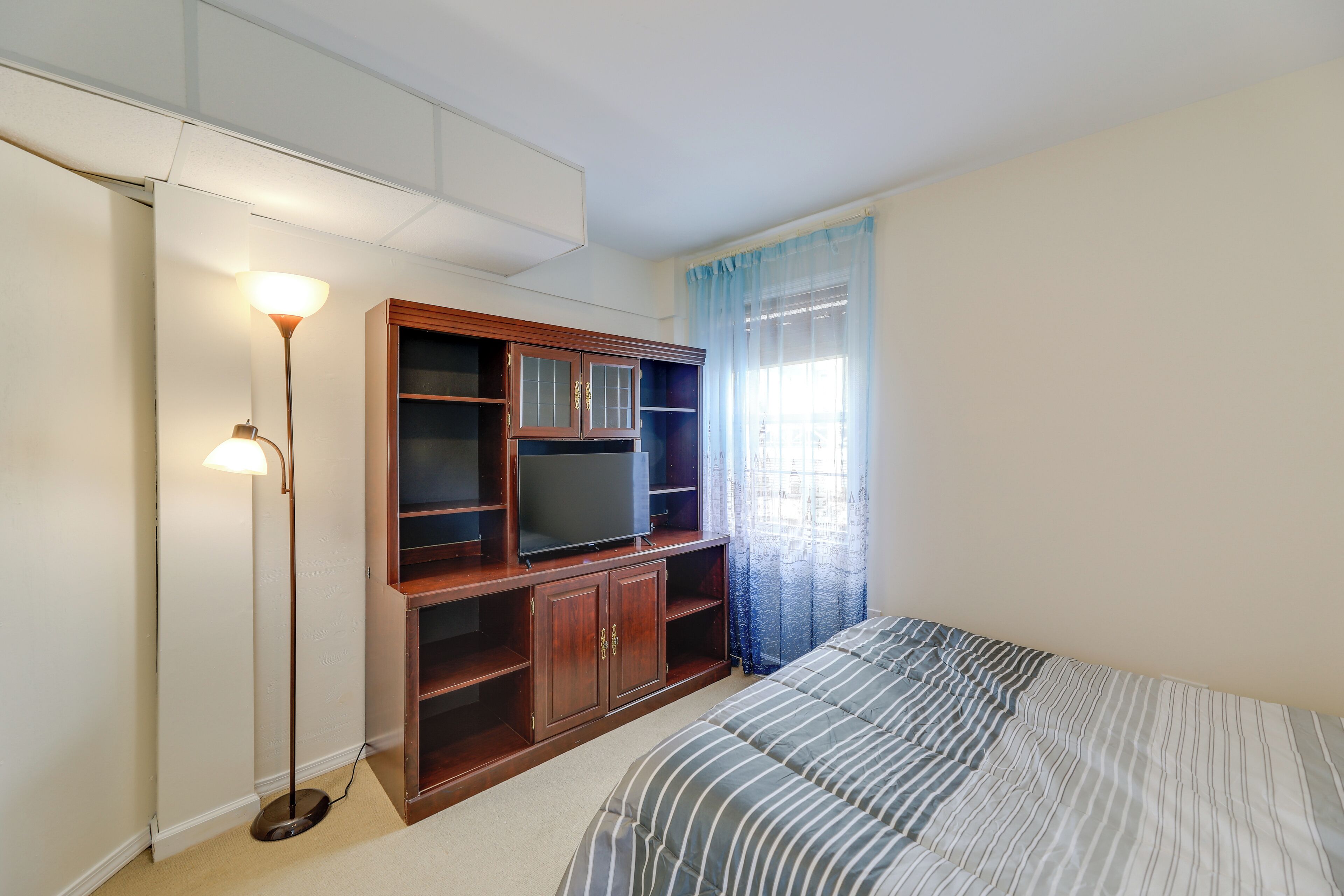 A bedroom with a bed, a TV on a wooden cabinet, a floor lamp, and a window with curtains.