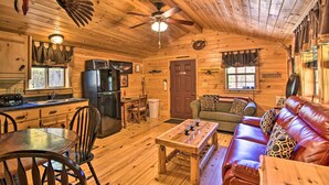 Interior de una cabaña con piso de madera, zona de comedor con sillas y mesa, sofá de cuero, ventilador de techo y cocina con refrigerador.