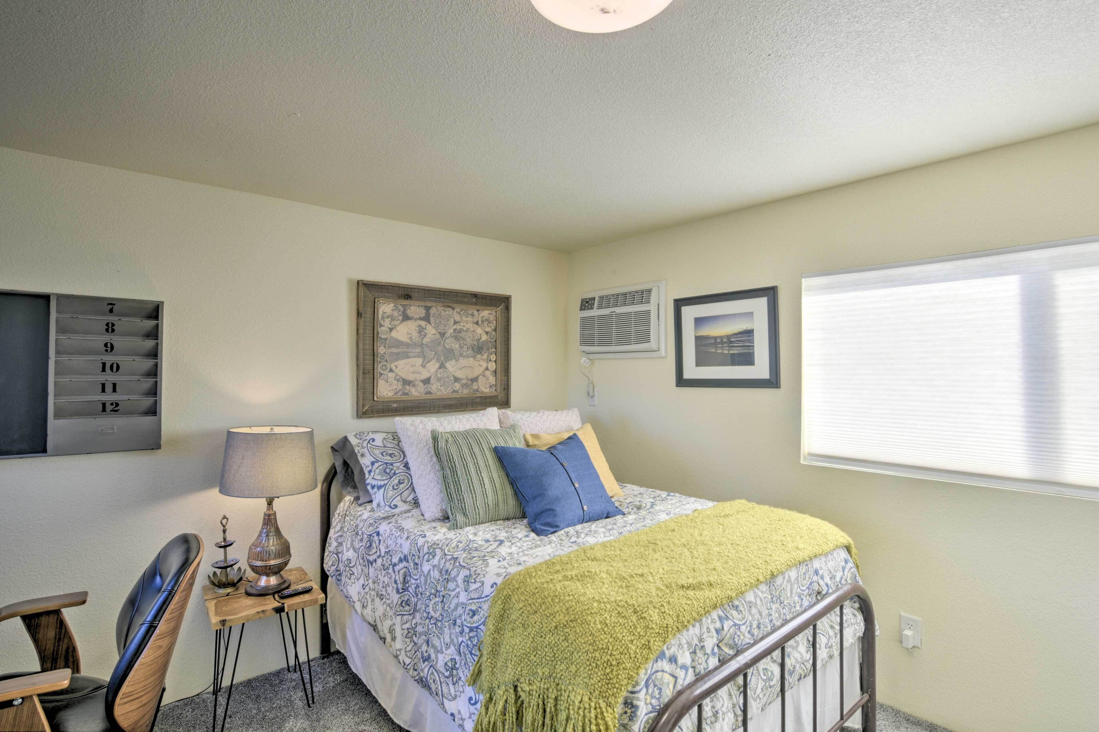A bedroom with a bed, a chair, a lamp, a small table, and a wall-mounted air conditioning unit.