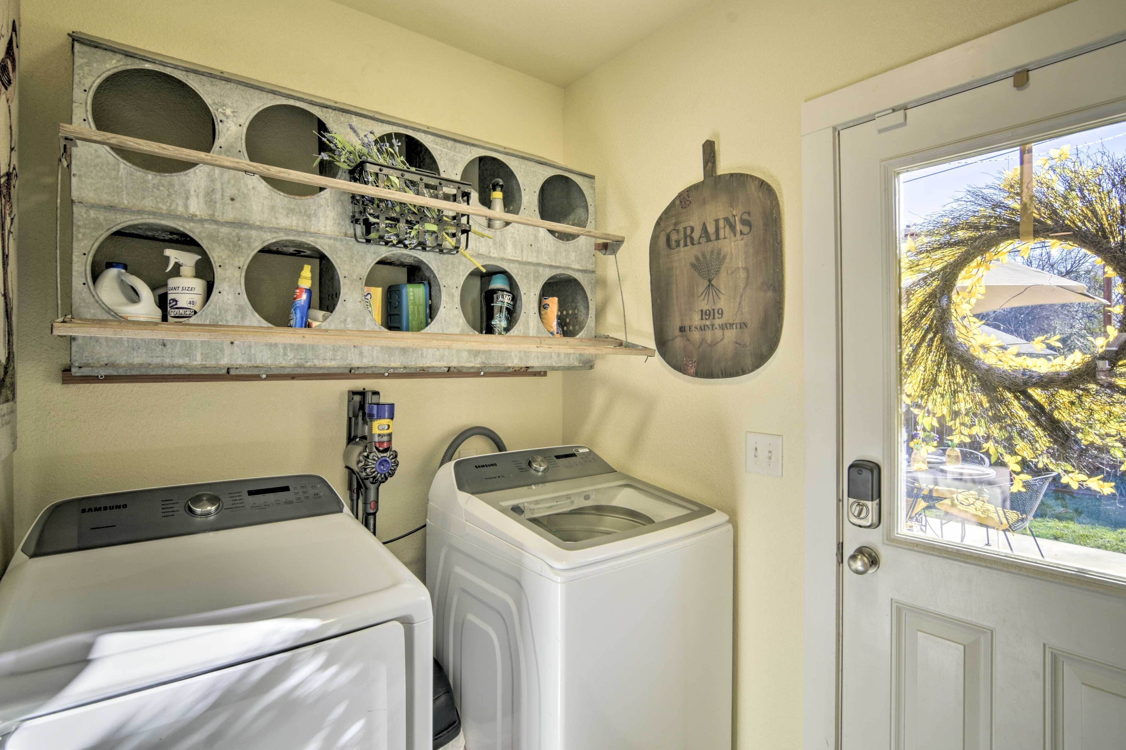 A laundry room with two white washing machines, a shelf with cleaning supplies, and a door with a view of trees.