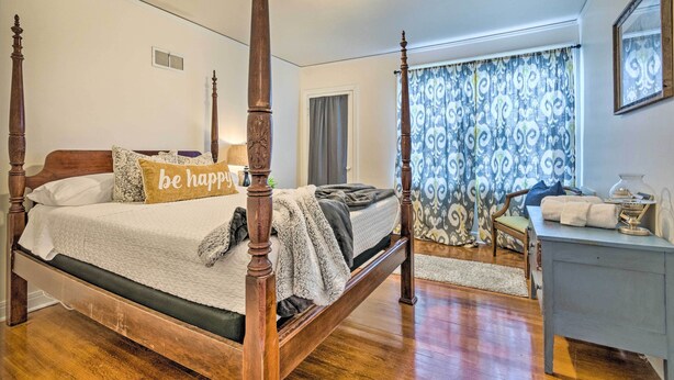 Un lit à baldaquin avec un coussin orné de la mention « soyez heureux », une table de chevet avec une lampe et une fenêtre agrémentée de rideaux à motifs.