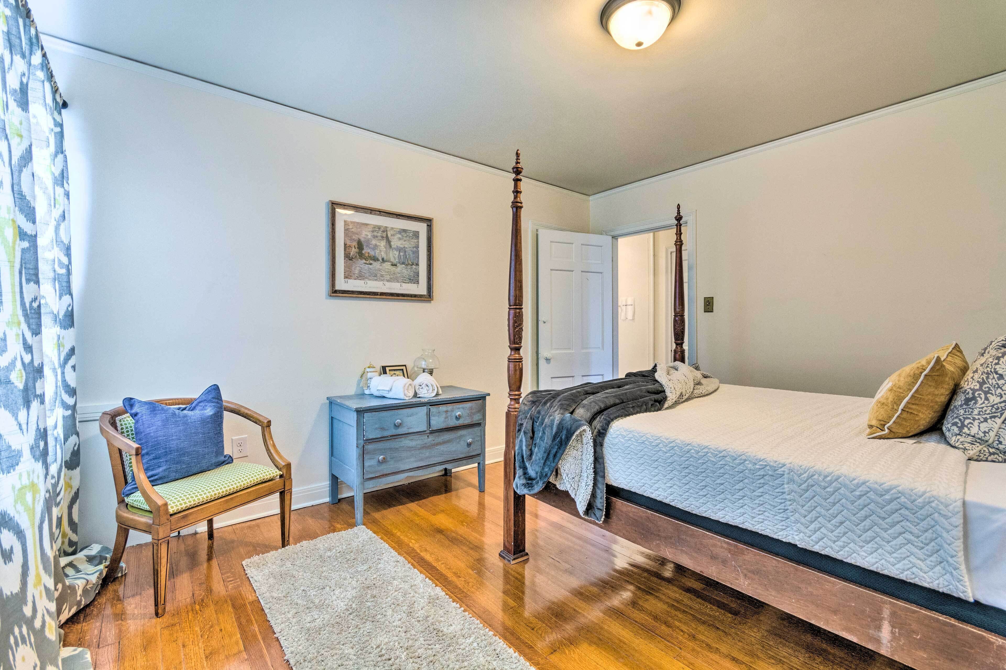 A bedroom with a four-poster bed, a wooden chair with a cushion, a dresser, and a framed picture on the wall.