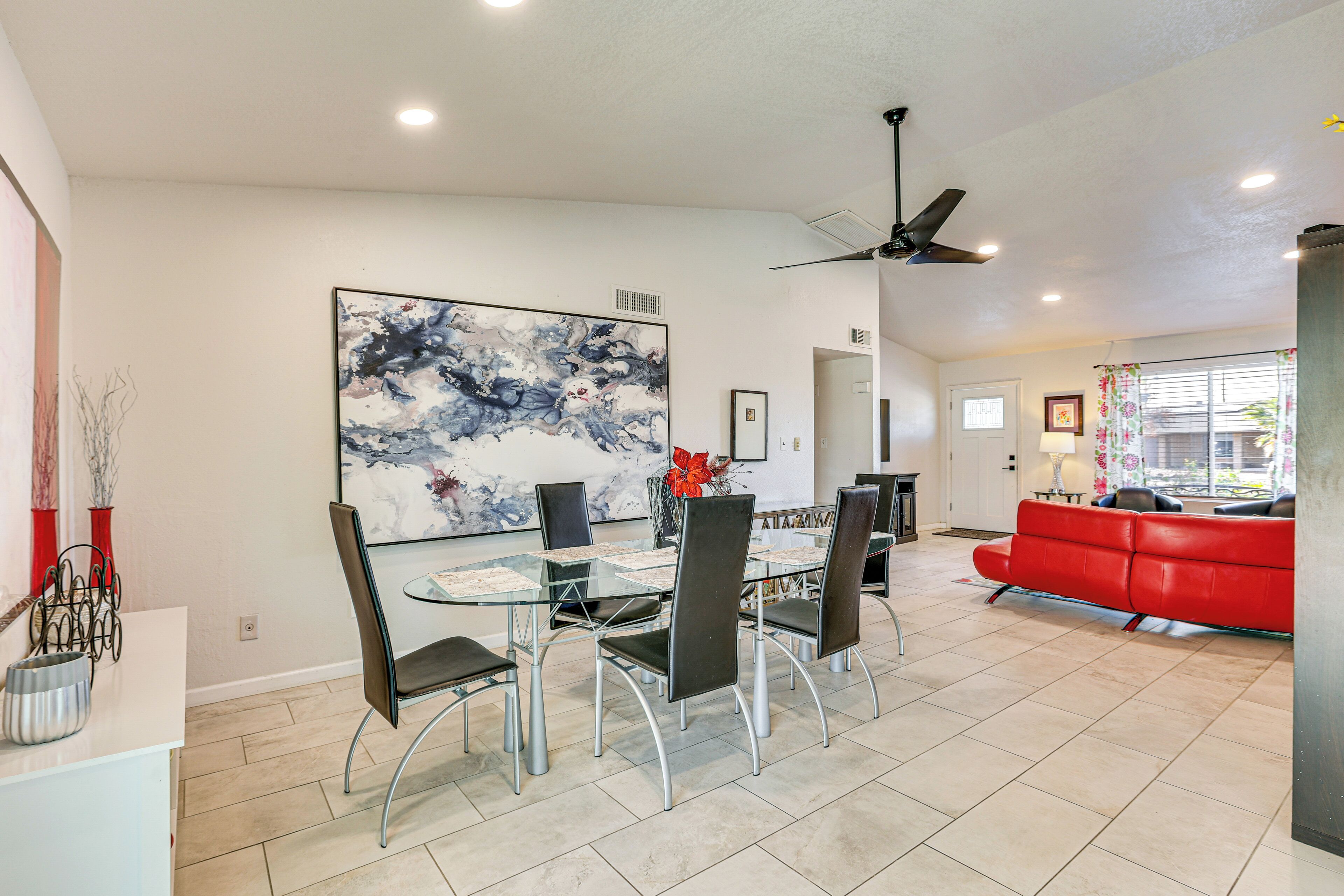 A modern dining area with a glass table and metal chairs, a large abstract painting on the wall, and a red sofa in the living room.