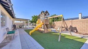 Un jardin avec un espace de jeux comprenant un toboggan et une balançoire, une terrasse avec des meubles d’extérieur et un mur en briques.