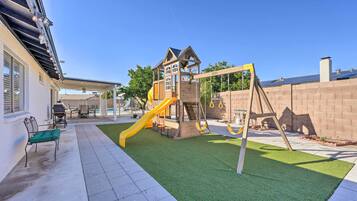 Un jardin avec un espace de jeux comprenant un toboggan et une balançoire, une terrasse avec des meubles d’extérieur et un mur en briques.