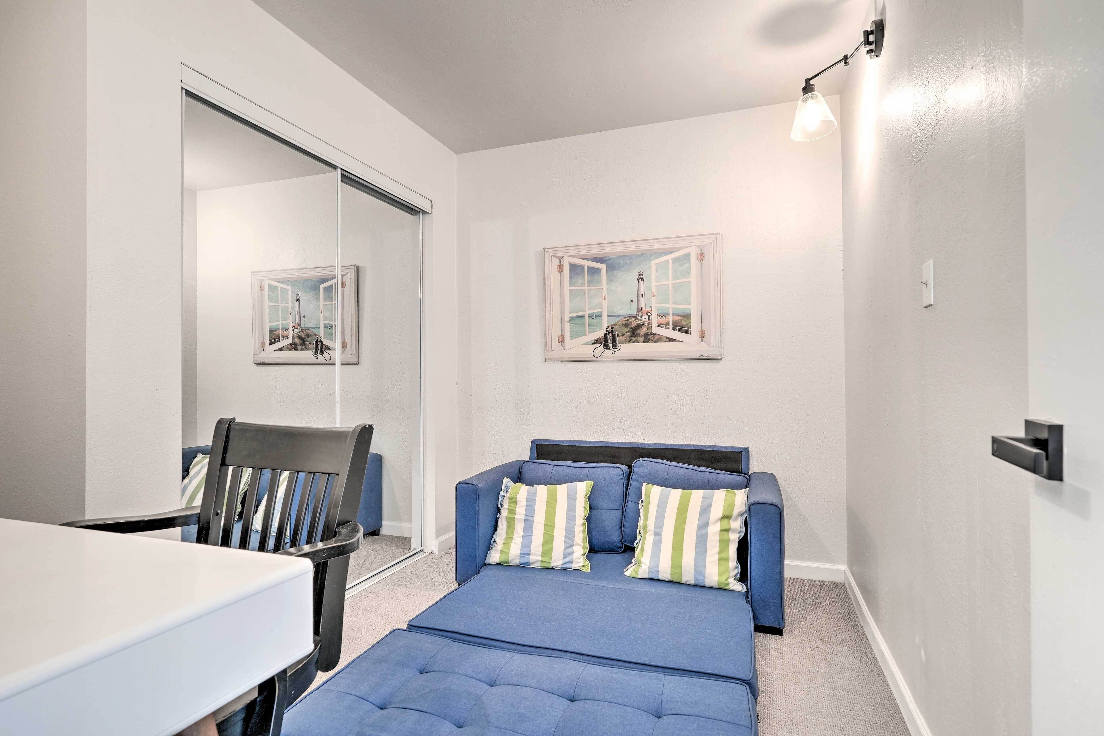 A room with a blue sofa, a dining table, and a mirror reflecting a painting of a lighthouse.