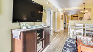 Un salon doté d’un téléviseur à écran plat fixé au mur, d’un meuble de rangement en bois, d’un coin repas avec une table et des chaises, et d’une cuisine équipée d’un four à micro-ondes.