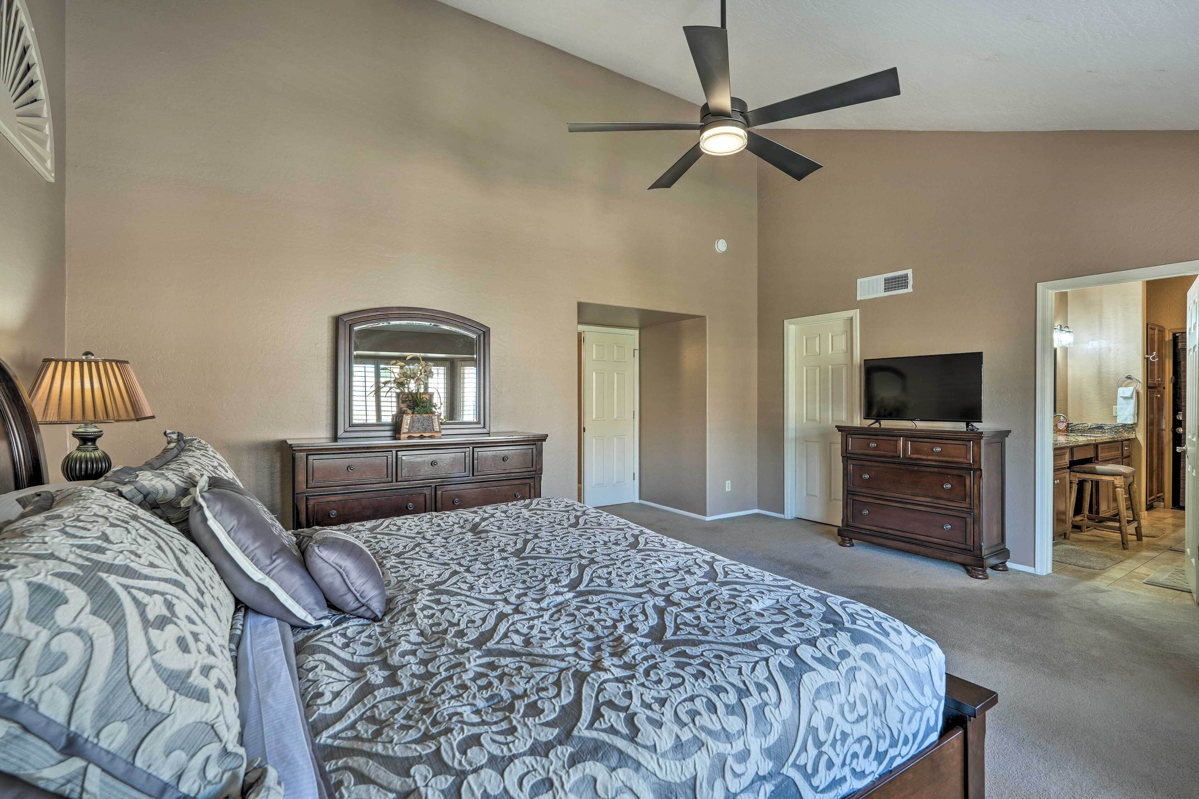 Une chambre avec un grand lit, un ventilateur de plafond, une commode, un téléviseur et une salle de bain visible à travers une porte ouverte.