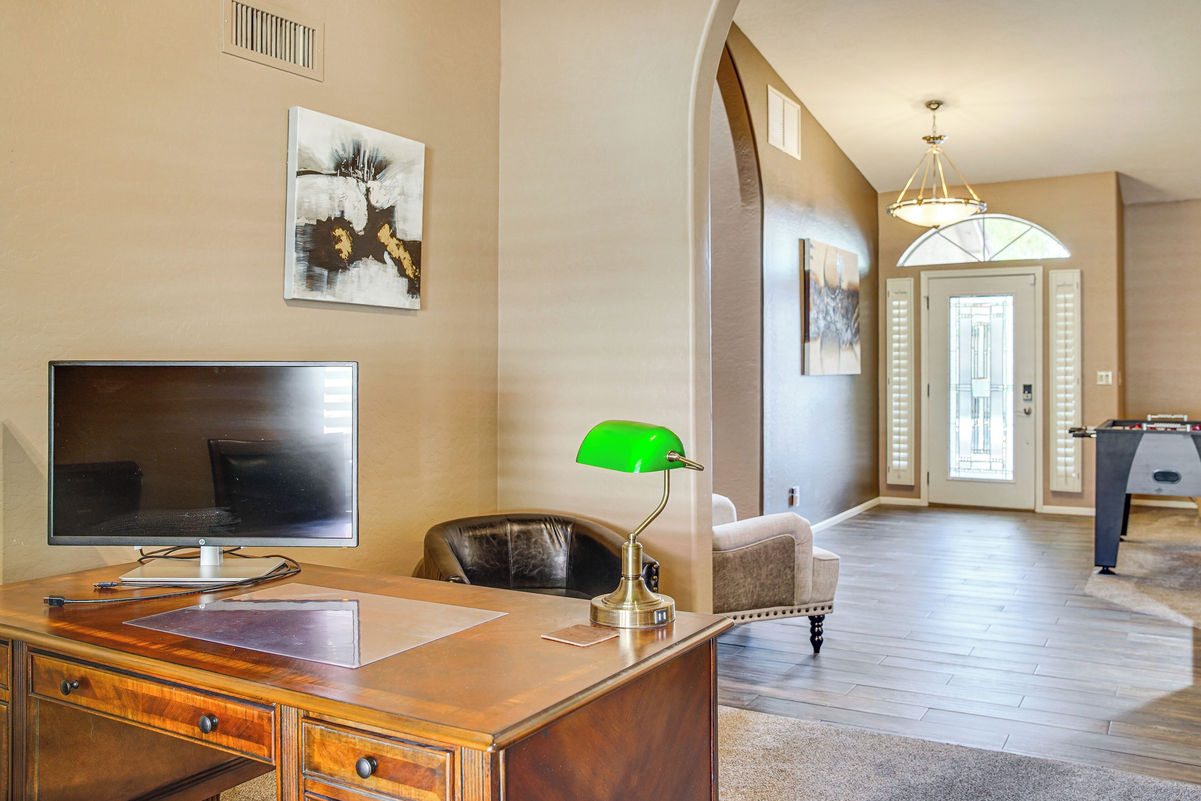A spacious room with a wooden desk, a flat-screen TV, a green desk lamp, and a leather chair. There is a door leading to another room with a foosball table.