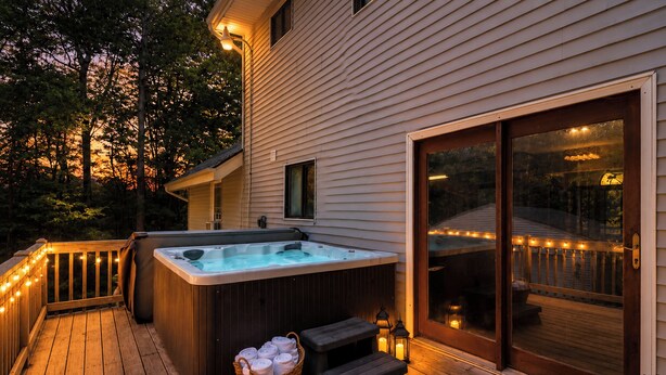 Jacuzzi en terraza de madera con una cesta de toallas y una mesa, cerca de una casa con puerta corrediza de vidrio.