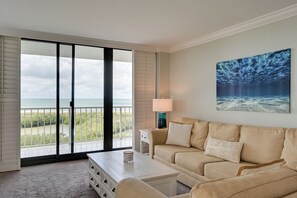 Een woonkamer met een beige bank, een salontafel en een schilderij van een onderwaterlandschap. Een schuifdeur leidt naar een balkon met uitzicht op het strand en de oceaan.