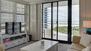 Un salon avec un téléviseur, une table basse et un canapé. Une grande fenêtre offre une vue sur un balcon et un immeuble.