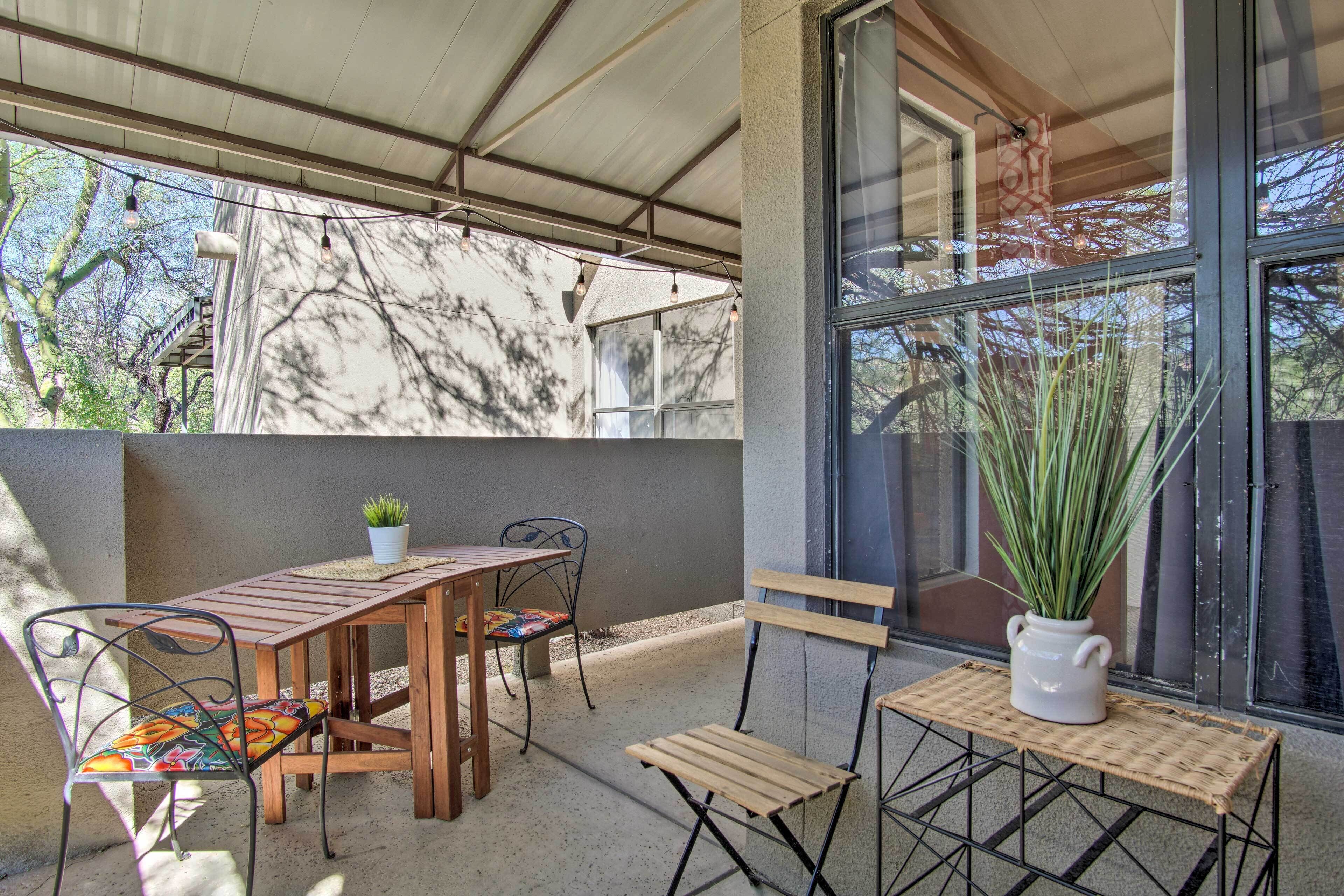 An outdoor patio with a wooden table and chairs, a potted plant, and a view of trees through large windows.