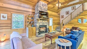 A cozy cabin interior with a stone fireplace, wooden floors, a sofa, and a staircase.