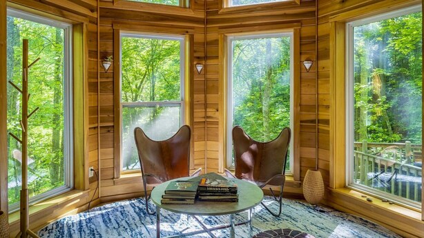 A cozy cabin interior with wooden walls, large windows offering a view of a forest, a round table with books, and two leather chairs.