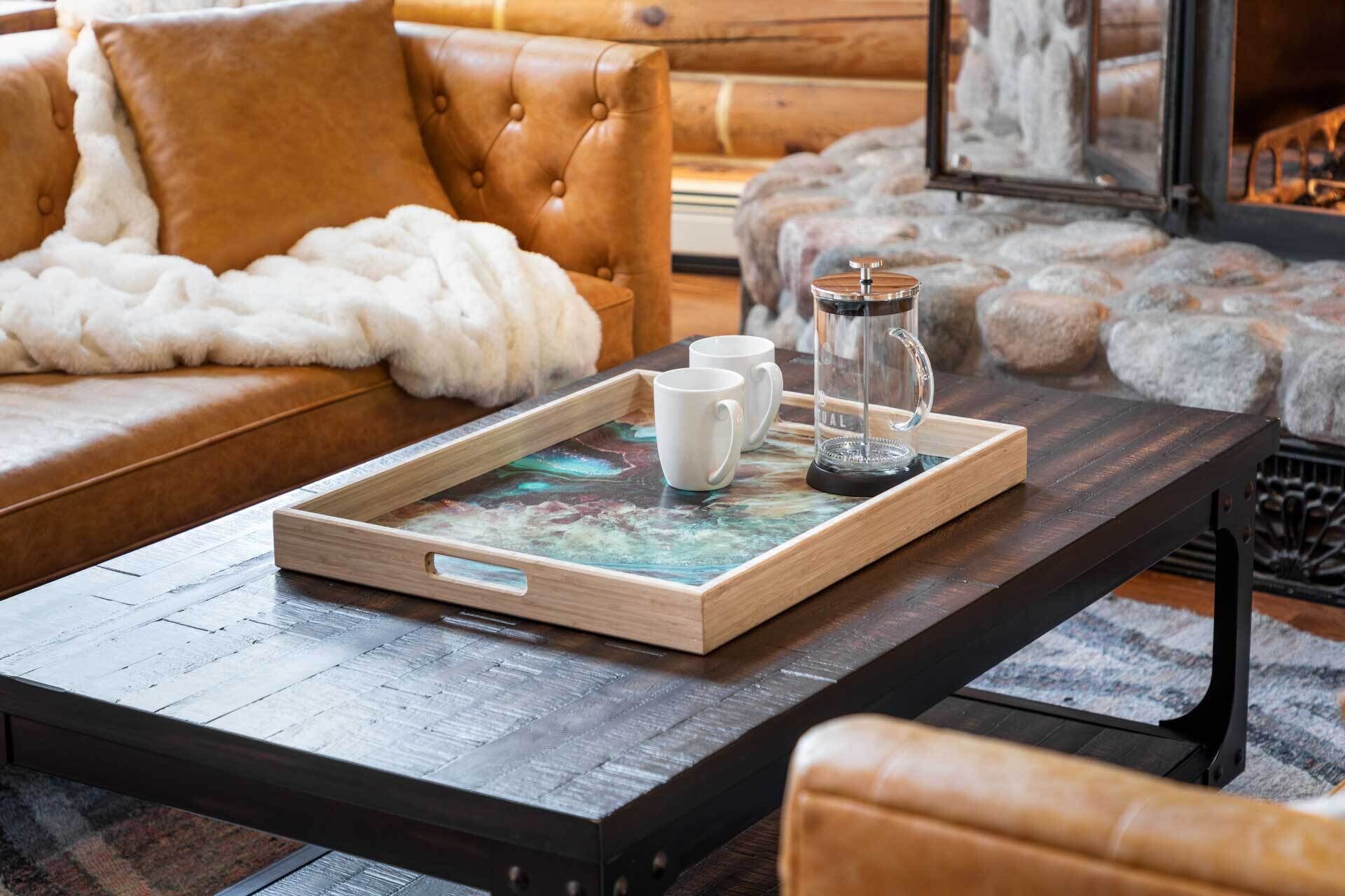 A wooden coffee table with a tray, two white mugs, and a glass carafe in a cozy living room with a leather sofa and a fur throw.
