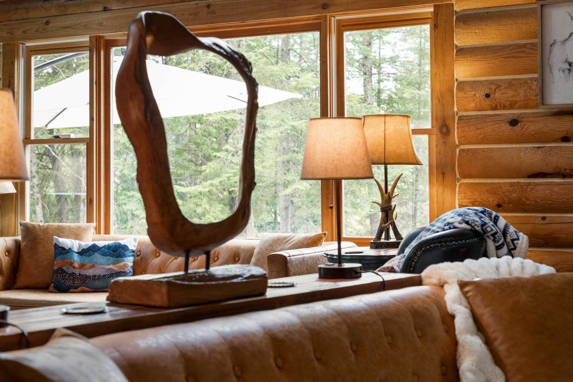 A cozy cabin interior with a large harp sculpture, a lamp, and a view of the forest through large windows.