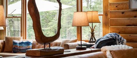 A cozy cabin interior with a large harp sculpture, a lamp, and a view of the forest through large windows.