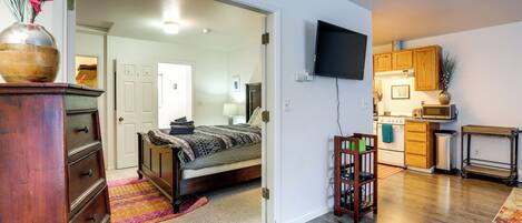 Un dormitorio con cama, cómoda y un jarrón de flores. Hay una zona de cocina con armarios y un microondas.