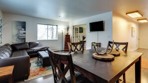 A modern living room with a leather sofa, wooden dining table, and chairs. There is a flat-screen TV mounted on the wall and a large window providing natural light. - Water-view Retreat! Steps to Coastal Trail & Dtwn (Anchorage)
